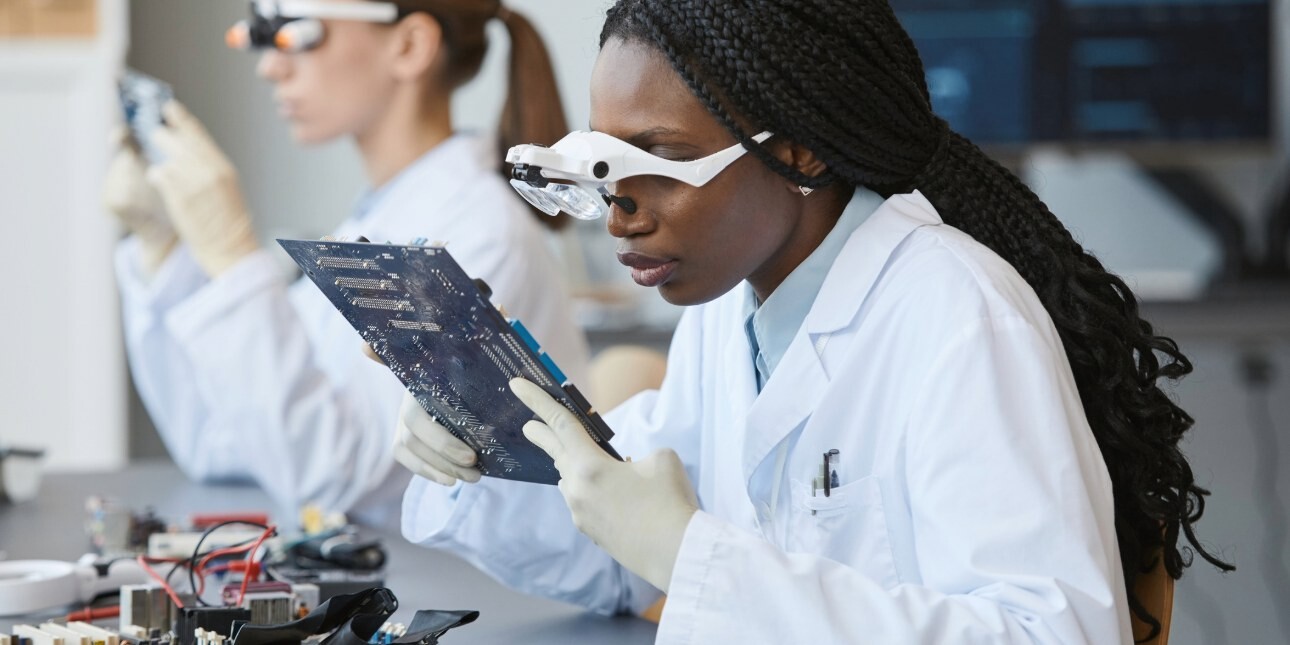 Two female scientists in a laboratory. The scientist in the foreground is examining a circuit board while wearing laboratory glasses. She wears a white lab coat.